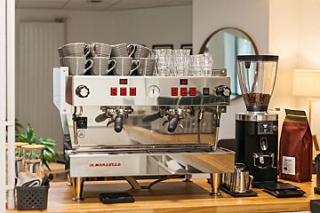 A professional La Marzocco espresso machine with stacked grey cups and clear glasses, alongside a coffee grinder and coffee bag on a wooden counter.