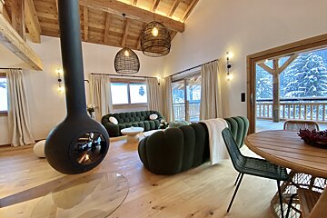 A modern, cozy cabin living room with a suspended fireplace, plush green sofas, and large windows overlooking a snowy forest landscape.