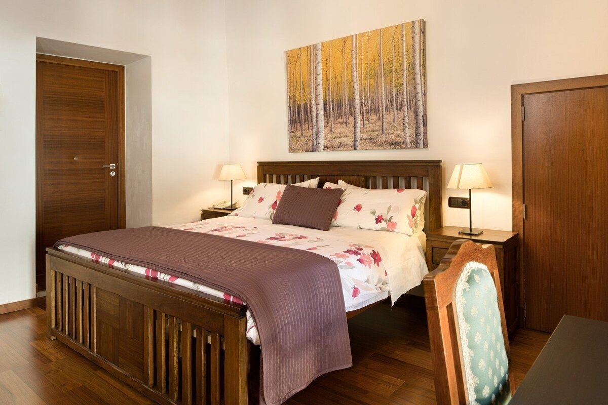 A cozy bedroom features a wooden bed with floral bedding, framed by wooden nightstands and lamps. A nature art piece hangs above, complemented by warm wooden floors and doors.