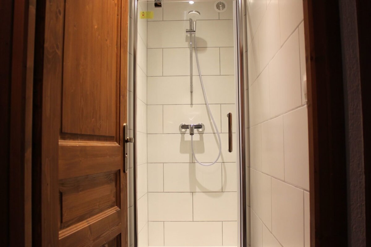 A modern shower stall with white rectangular tiles, a chrome showerhead and mixer, enclosed by a glass door. A wooden door is partially open on the left.