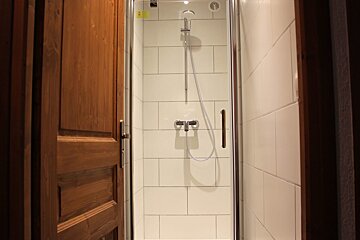A modern shower stall with white rectangular tiles, a chrome showerhead and mixer, enclosed by a glass door. A wooden door is partially open on the left.