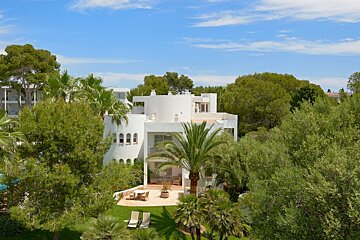 A white modern villa with a patio & loungers, enveloped by lush green trees & palms under a clear blue sky.