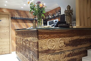 An ornate carved wooden bar with flowers, spirits, and a statue. Warm interior with a restroom door visible, suggesting a restaurant or bar.