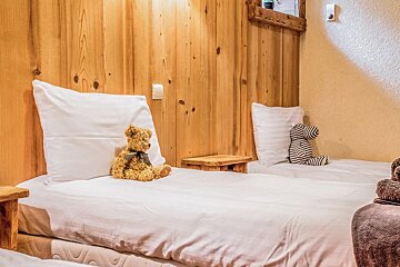 Two single beds with white bedding, a teddy bear, and a striped stuffed animal are set against a warm wooden wall in a cozy room.