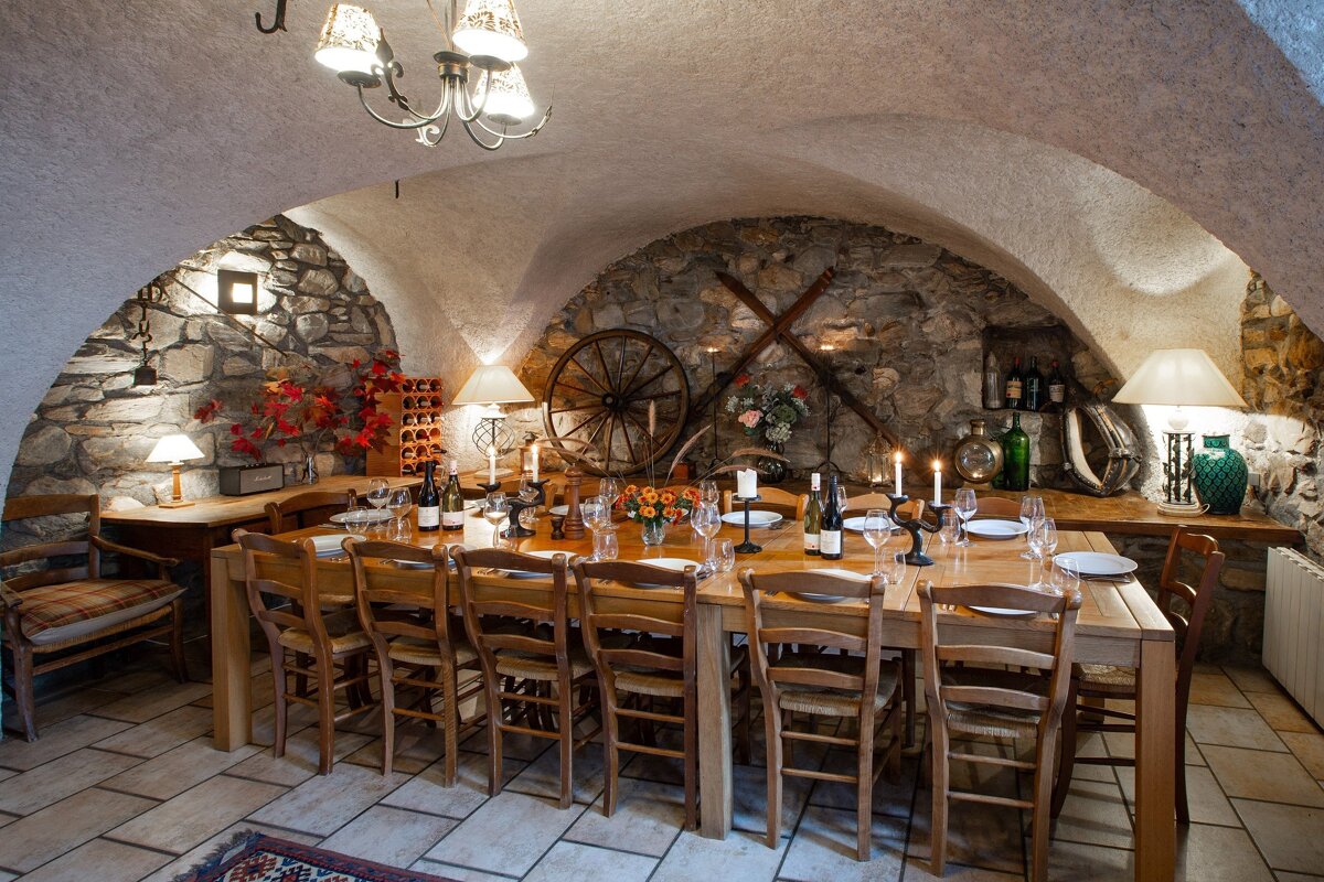 A rustic vaulted dining room features a long wooden table set with dinnerware, wine, and candles. Stone walls are adorned with a wagon wheel and lanterns.