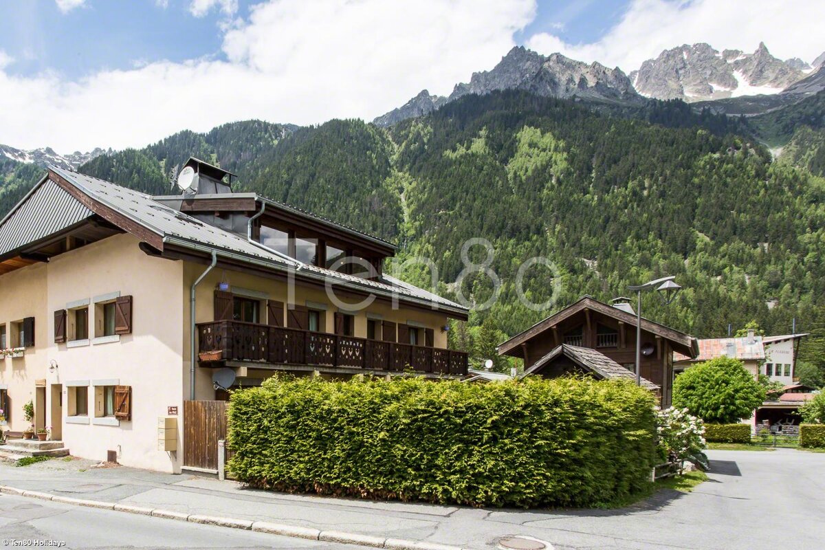 A house with a balcony is surrounded by a hedge and mountains in the background