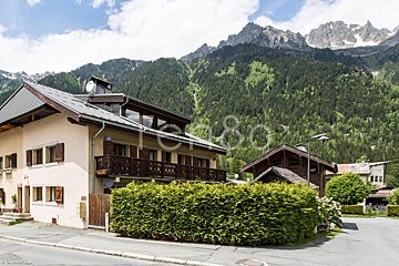 A house with a balcony is surrounded by a hedge and mountains in the background