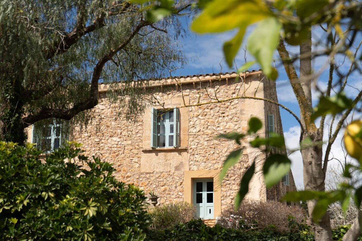 A rustic stone house with blue shutters and a door, peeking through green trees under a clear blue sky.