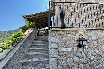 A stone building with outdoor steps leading to a covered terrace. Mountains are visible in the background under a blue sky. A lamp and 'MESTRAL' sign are on the wall.