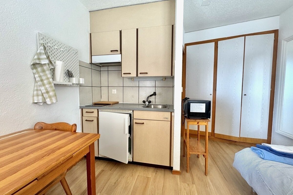 A small studio apartment featuring a kitchenette with a sink and fridge, a wooden dining table, a wardrobe, and a bed with striped bedding.