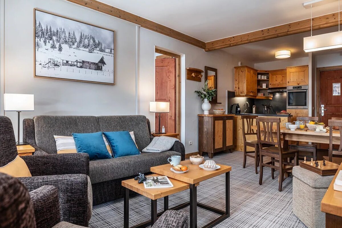Cozy cabin-style living area with a gray sofa, blue pillows, wooden coffee tables, a dining set, and an open kitchen.