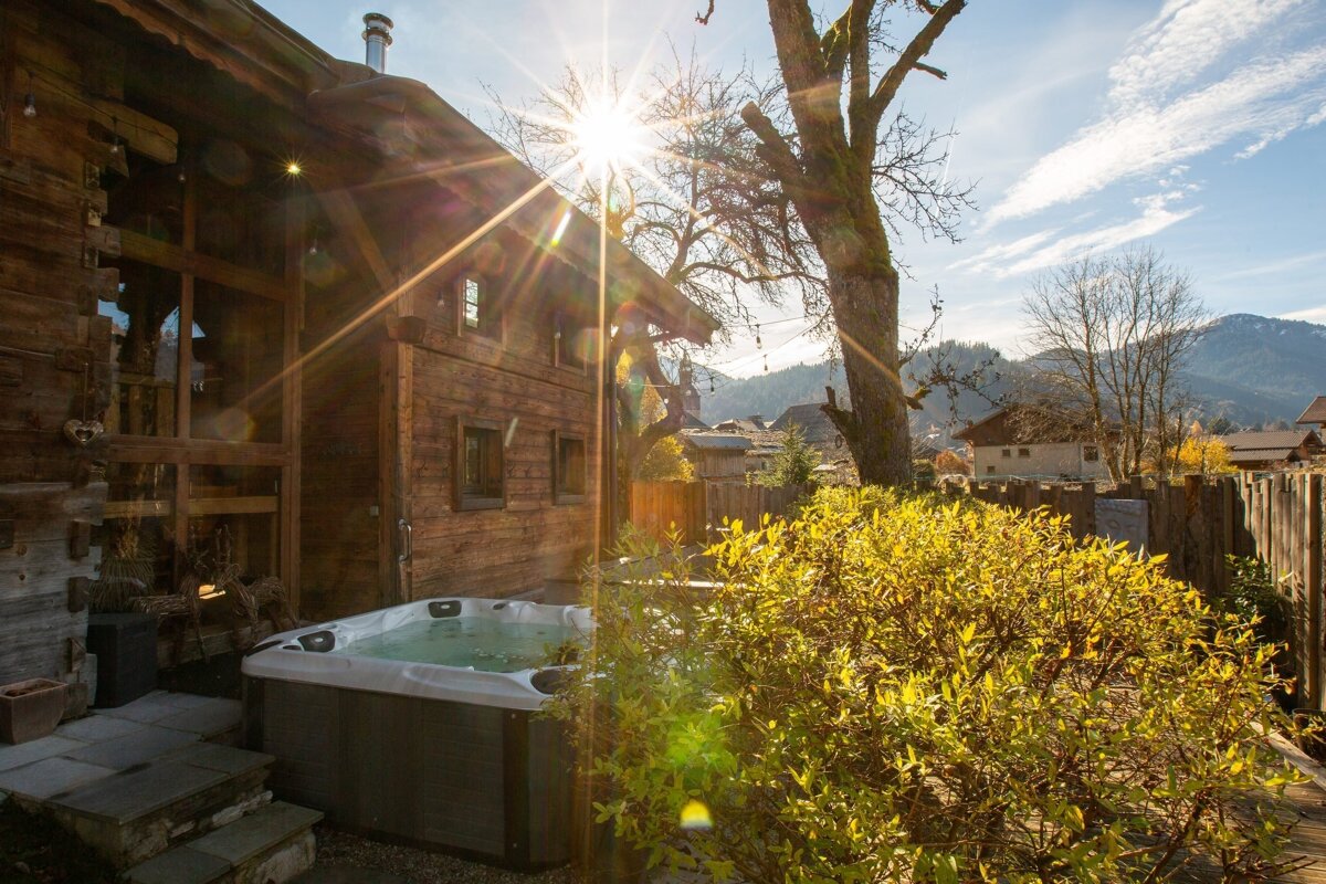 A cozy wooden cabin featuring an outdoor hot tub, with bright sunshine and a scenic mountain backdrop.