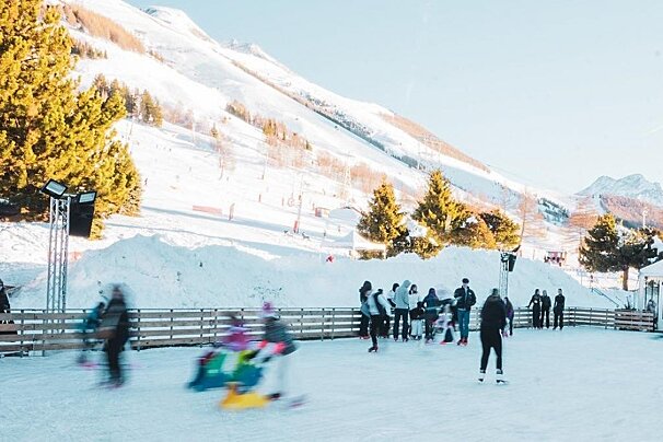 People ice skating outdoors on a sunny day with snow-covered mountains and pine trees in the background. Some skaters are blurred, indicating motion.