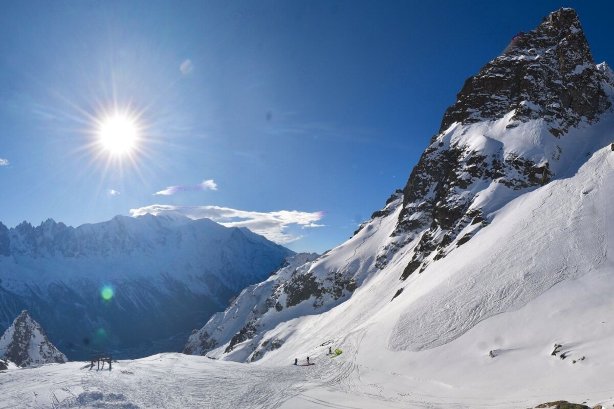 A dazzling sun illuminates vast snow-covered mountains under a brilliant blue sky, with tiny figures on the snowy slopes.