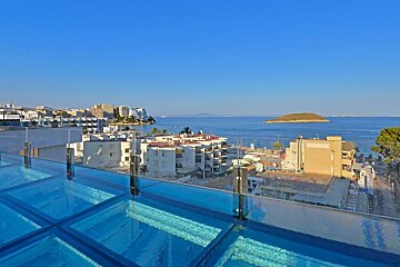 Rooftop pool with glass panels overlooks a sunny coastal town, vibrant blue sea, and a small island in the distance.