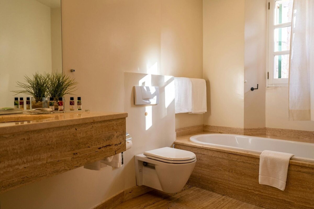 A bright, modern bathroom with a floating toilet, marble vanity, bathtub, and natural light from a window, featuring neutral tones.