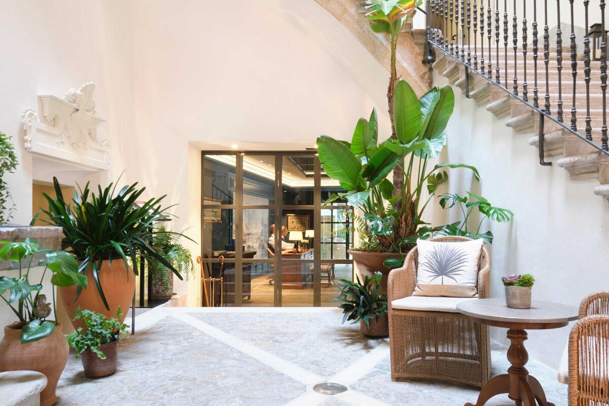 An inviting indoor atrium features abundant potted tropical plants, a grand stone staircase, wicker chairs, and glass doors revealing a room beyond.