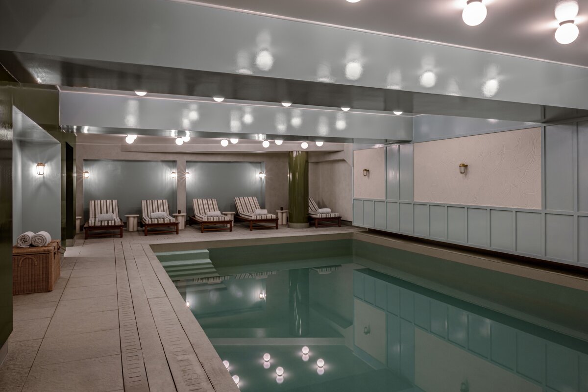 A tranquil indoor pool area featuring a light blue/grey color scheme, striped lounge chairs, and ceiling lights reflecting in the calm water.