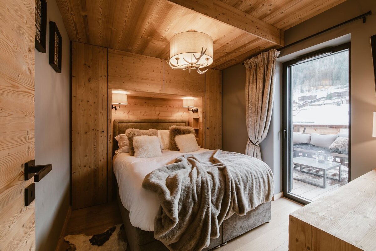 A cozy, chalet-style bedroom with warm wood paneling, a plush bed, and a balcony offering a snowy mountain view.