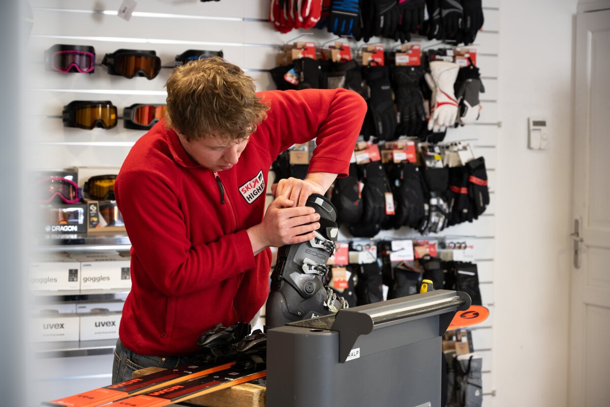 A man in a red fleece adjusts a ski boot in a shop, surrounded by ski goggles and gloves on display.