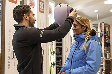 A man wearing a skiset jacket helps a woman put on a helmet