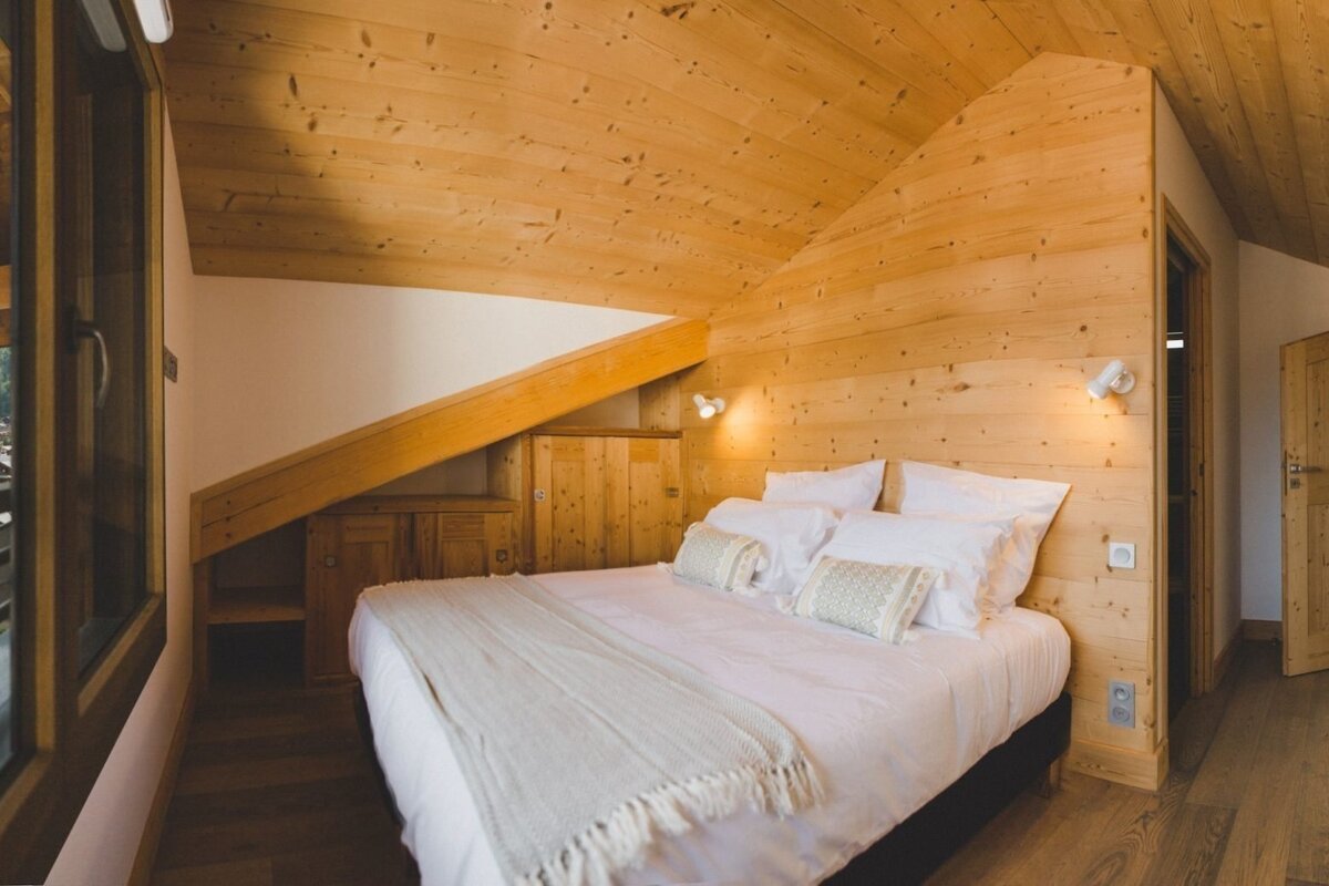 A cozy, rustic attic bedroom featuring extensive light wood paneling, a sloped ceiling, and a large bed with white bedding.