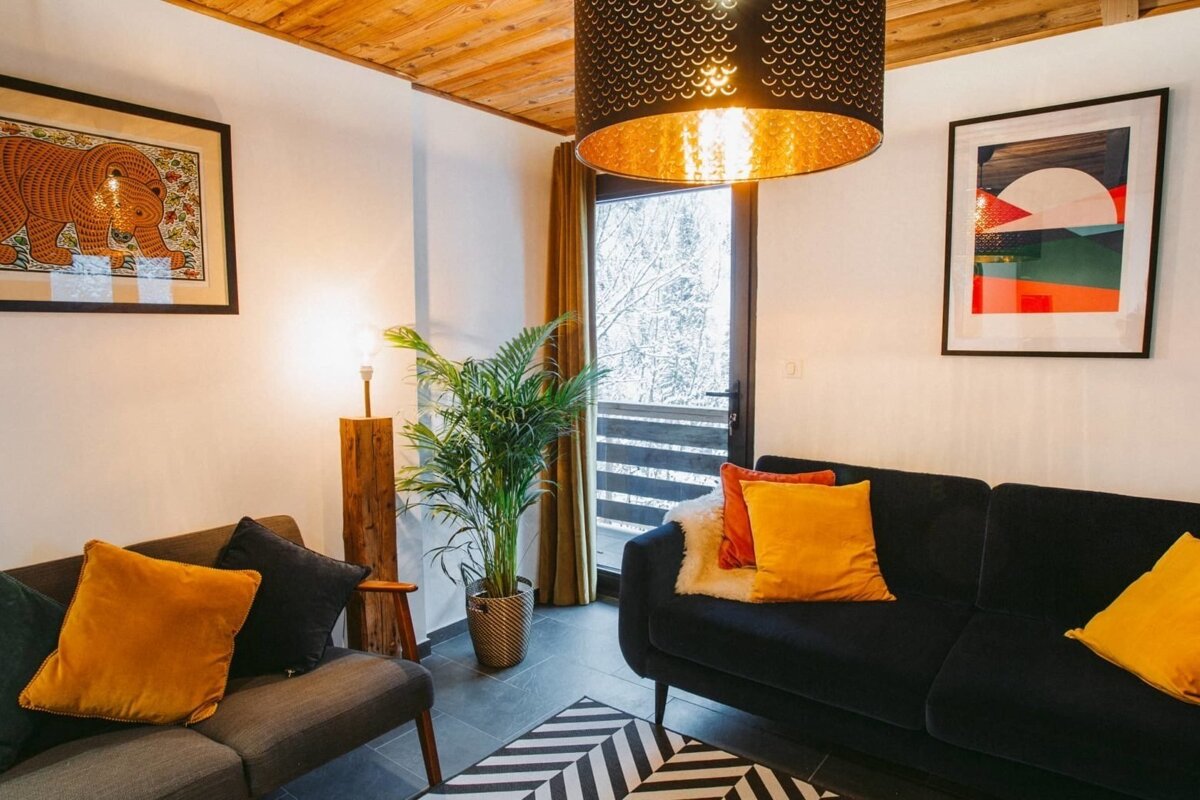 A cozy living room with dark sofas, warm orange accents, wooden ceiling, and a view of snowy trees. Artwork adorns the walls.