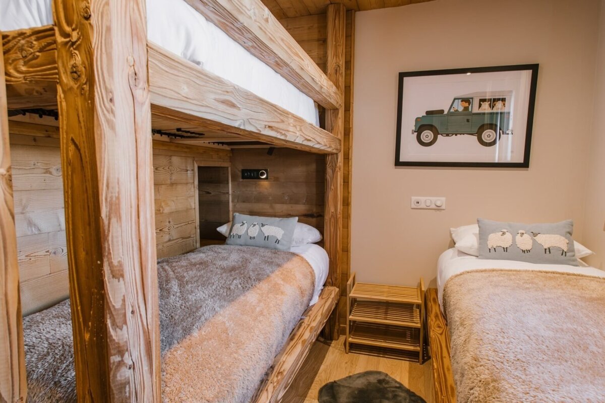 Cozy, rustic bedroom featuring a sturdy wooden bunk bed, a single bed with sheep pillows, and a framed car illustration on the wall.