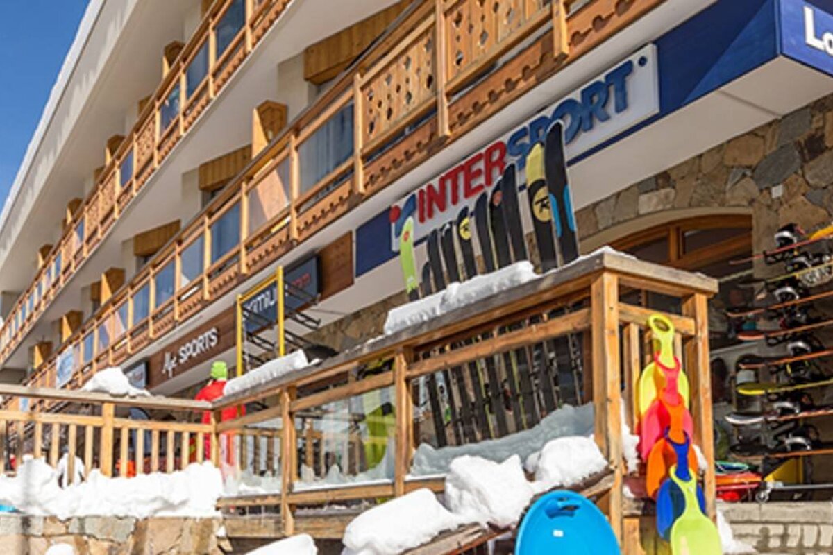 A snow-covered ski shop (Intersport) at a resort building, featuring skis, snowboards, and sleds on display under a bright sky.