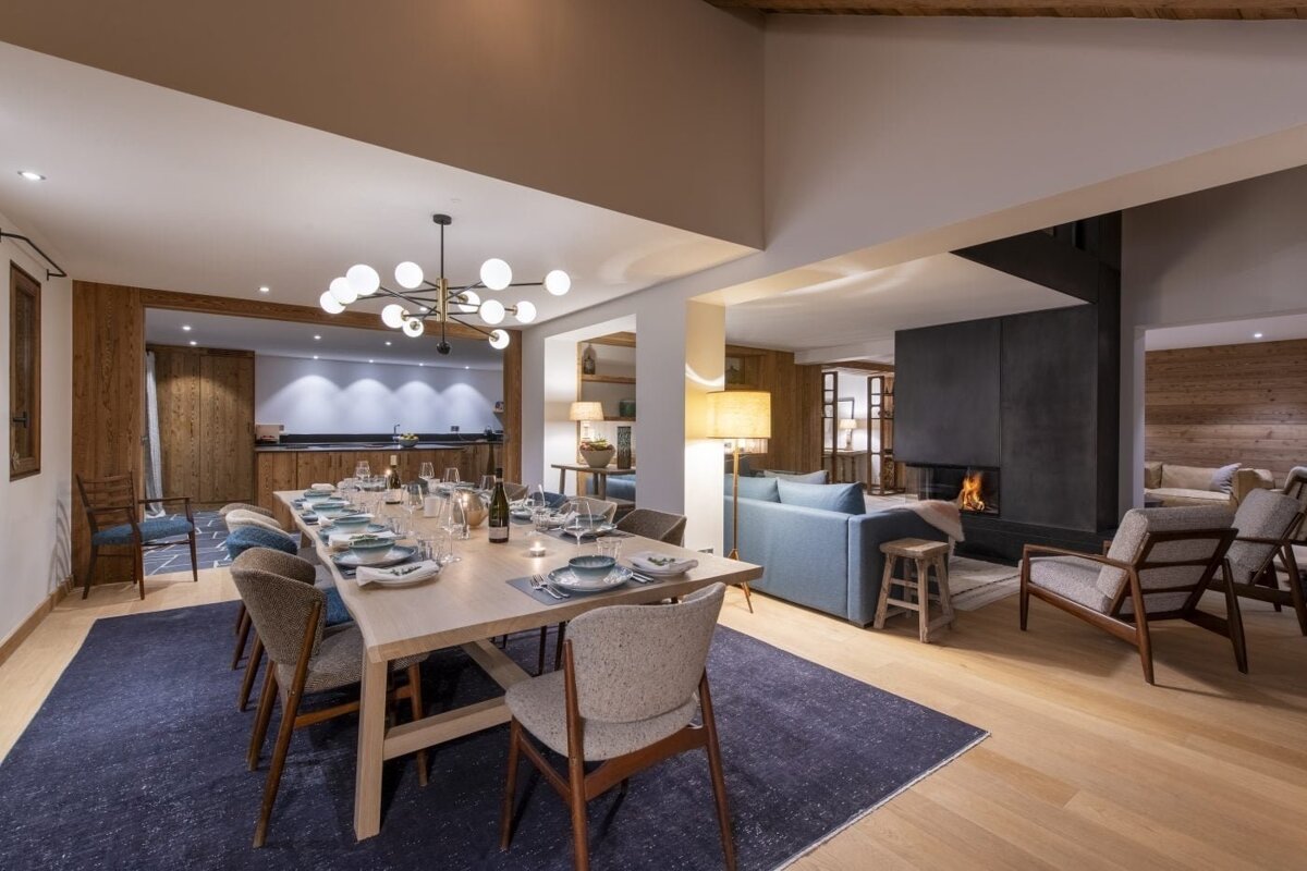 A spacious, modern open-plan living and dining area features a set table, contemporary chandelier, cozy fireplace, blue sofa, and wooden accents.