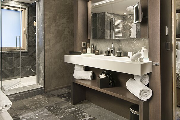 A modern, luxurious bathroom with dark marble flooring and walls. It features a floating white double vanity, large mirror, and glass-enclosed walk-in shower.