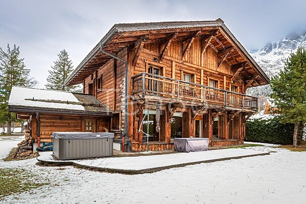 A rustic wooden chalet with snow-covered ground, two hot tubs, and snowy mountains in the background.