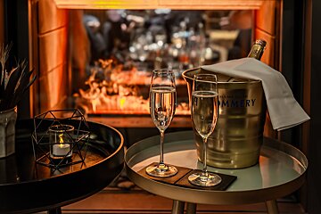 Two champagne flutes and a bottle in an ice bucket sit on tables, with a warm, flickering fireplace in the background, creating a cozy atmosphere.