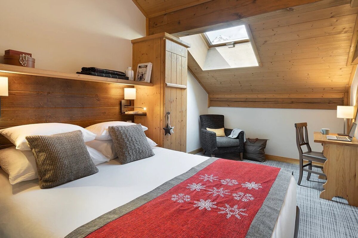 Cozy attic bedroom with wooden paneling, a skylight, a bed with a red snowflake runner, an armchair, and a small study desk.