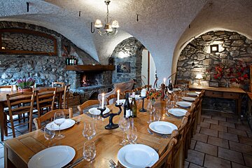 A cozy, rustic dining room features stone walls, an arched ceiling, and a long wooden table set with plates, glasses, and candles, all warmed by a fireplace.