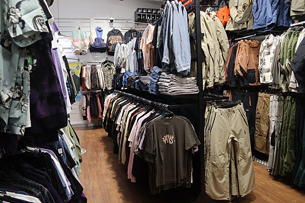 A well-stocked clothing store interior with racks and shelves displaying a wide variety of shirts, jackets, pants, and hoodies on a wooden floor.