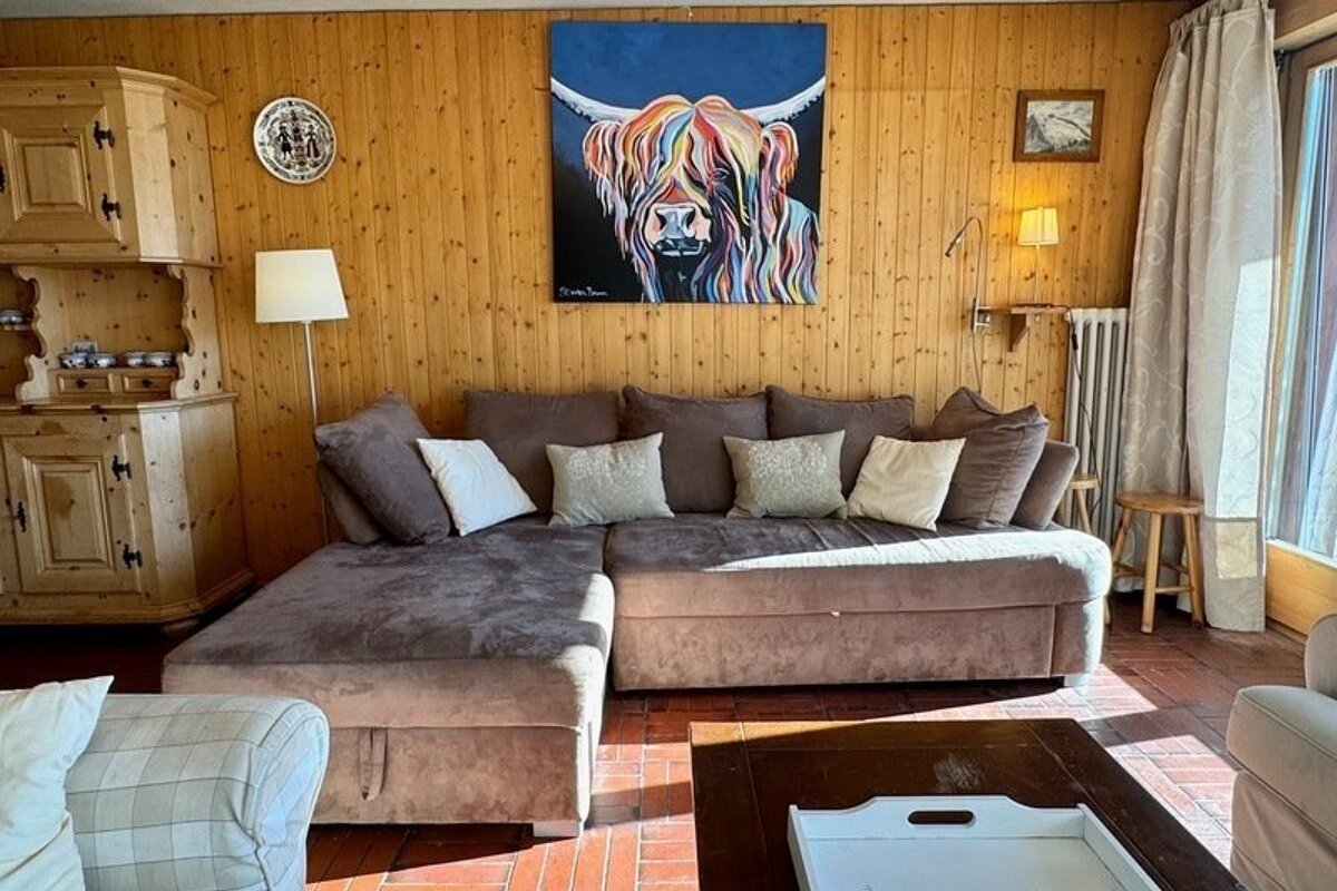 A cozy wooden living room featuring a brown sectional sofa, a coffee table, and a colorful highland cow painting on the wall.
