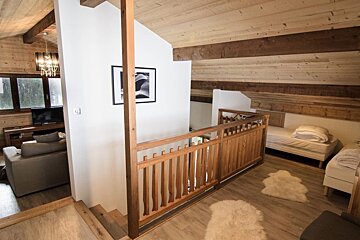 Cozy wood-paneled chalet interior with a split-level design. A wooden railing separates an upper bedroom area from a lower living room with a sofa and TV.