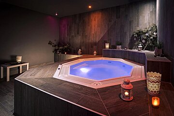 A tranquil spa room features an octagonal hot tub with glowing blue water, surrounded by dark wood-paneled surfaces and ambient candlelight, creating a relaxing atmosphere.