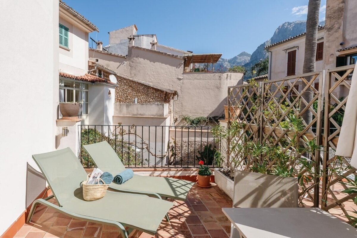Sunny terrace with two loungers on terracotta tiles, surrounded by old buildings and distant mountains under a blue sky.