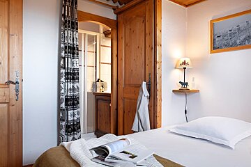 Cozy alpine-style bedroom with warm wood doors, white bedding, an open magazine, and a bedside lamp, leading to an ensuite bathroom.