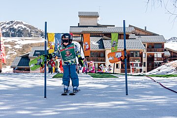 A child in ski gear stands on a snowy slope, framed by ski poles and cartoon flags, with buildings and mountains behind.