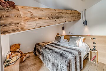 A cozy, rustic-modern bedroom featuring a large wooden beam, slanted ceiling, faux fur blanket on the bed, and hardwood floors. A teddy bear sits nearby.