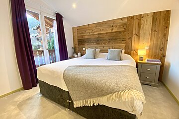 A cozy bedroom featuring a wooden headboard, large bed with a throw blanket, and plum curtains framing a balcony view of mountain chalets.