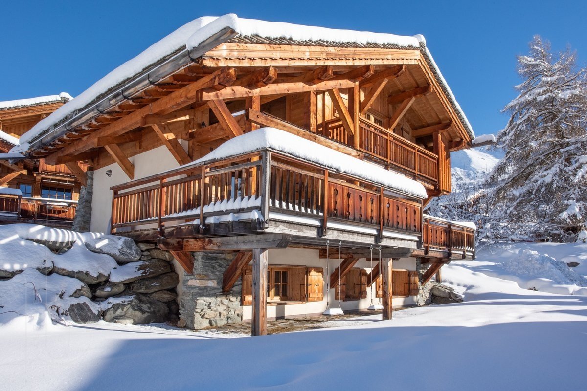 A rustic wooden chalet with balconies is covered in deep snow under a clear blue sky, set amidst a picturesque winter landscape with snowy trees.