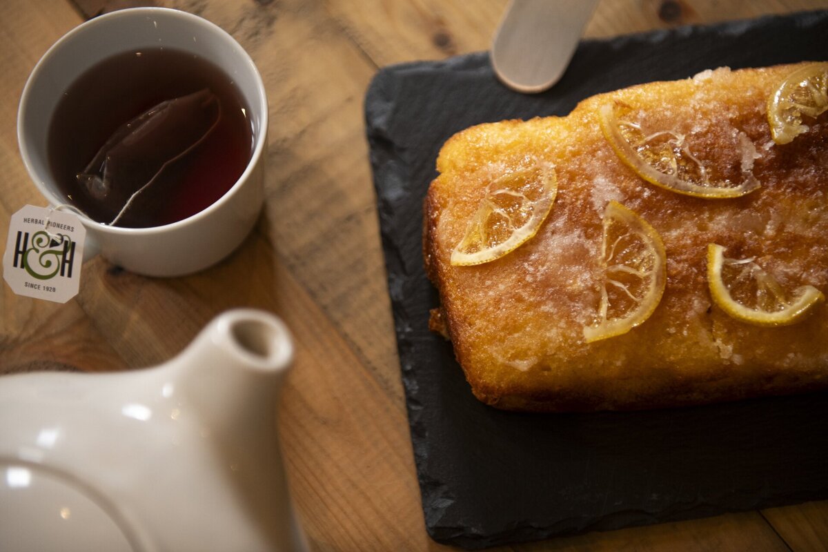 A cup of tea with a teabag sits beside a lemon loaf cake topped with sugared lemon slices, served on a slate board and wooden table.