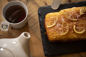 A cup of tea with a teabag sits beside a lemon loaf cake topped with sugared lemon slices, served on a slate board and wooden table.
