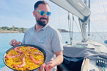 A man on a sailboat holding a pan of shrimp paella