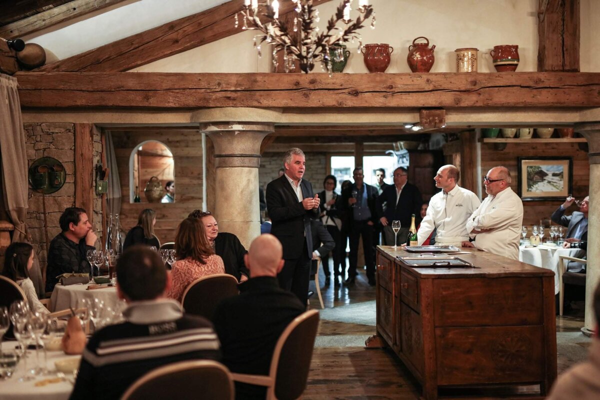 A man speaks to an audience in a rustic restaurant, with two chefs standing by a wooden counter. Guests are seated at tables.