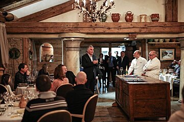 A man speaks to an audience in a rustic restaurant, with two chefs standing by a wooden counter. Guests are seated at tables.